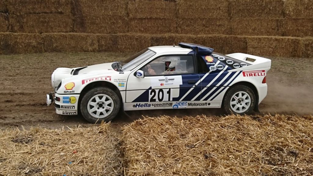 A Ford RS200 rally car driving between haybales, kicking up a dust plume.